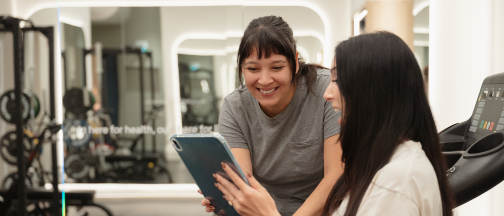 Two women in a gym looking at a tablet together, one smiling while the other views the screen